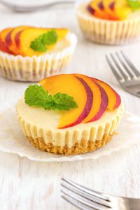 Mini cheesecakes topped with sliced peaches and a mint sprig on a white table, with silver forks nearby.