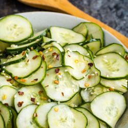 A bowl of sliced cucumbers topped with red pepper flakes and herbs, with a wooden spoon in the background.