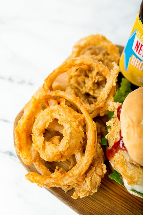 Close-up of crispy golden onion rings, a bottle of sauce, and part of a burger with lettuce, tomato, and cheese on a wooden board.