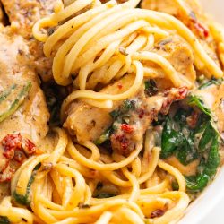 A close-up of a fork twirling spaghetti with pieces of chicken, spinach, and sun-dried tomatoes in a creamy sauce.