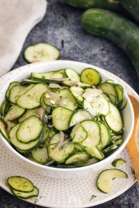 A white bowl filled with cucumber salad, featuring thinly sliced cucumbers and onions, garnished with herbs. Sliced cucumbers are scattered on the surface nearby along with whole cucumbers in the background.