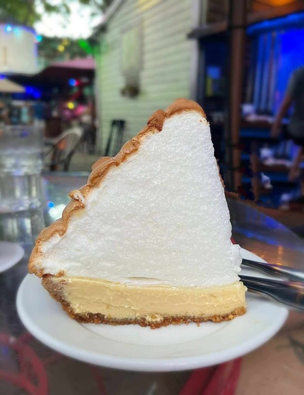A large slice of lemon meringue pie with a golden crust and tall, fluffy meringue topping sits on a white plate at an outdoor café table, reminiscent of some of the Best Key Lime Pies enjoyed in sunny coastal cafes.