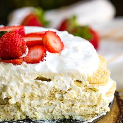 Close-up of a layered dessert with white cream, crushed cookies, and sliced strawberries on top, displayed on a wooden surface.