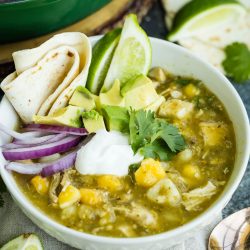 A bowl of green chili stew with chicken, potatoes, avocado slices, lime wedges, sour cream, onion, and flour tortillas beside it.