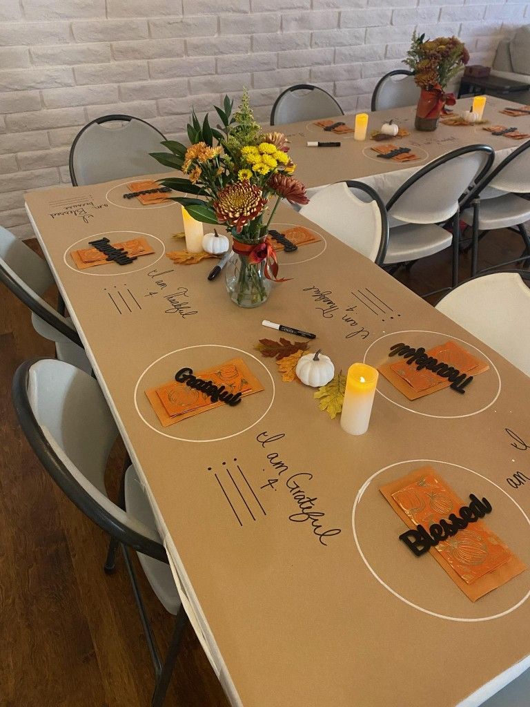 A table set for a fall-themed gathering with a brown paper table cover, autumn flowers, candles, white pumpkins, and place settings labeled "Grateful," "Thankful," and "Blessed"—perfect inspiration for your Thanksgiving Dinner Menu and Recipes.