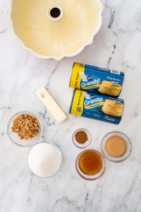 Ingredients for baking: bundt pan, two cans of flaky biscuits, butter, walnuts, sugar, cinnamon, vanilla extract, and brown sugar on a marble surface.