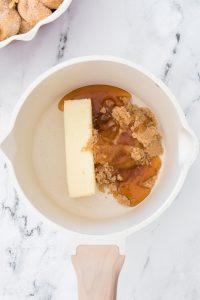 Stick of butter, brown sugar, and honey in a white saucepan on a marble surface.