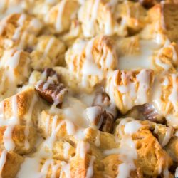 Close-up of a baked pastry dessert with a golden-brown surface, drizzled with white icing and pieces of chocolate throughout, in a glass baking dish.