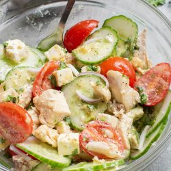A glass bowl of salad with chicken, cucumber slices, cherry tomatoes, feta cheese, onions, and herbs, with a spoon inside.