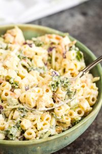 A close-up of a creamy pasta salad in a bowl, featuring macaroni, chopped greens, and shredded vegetables being served with a metal spoon.