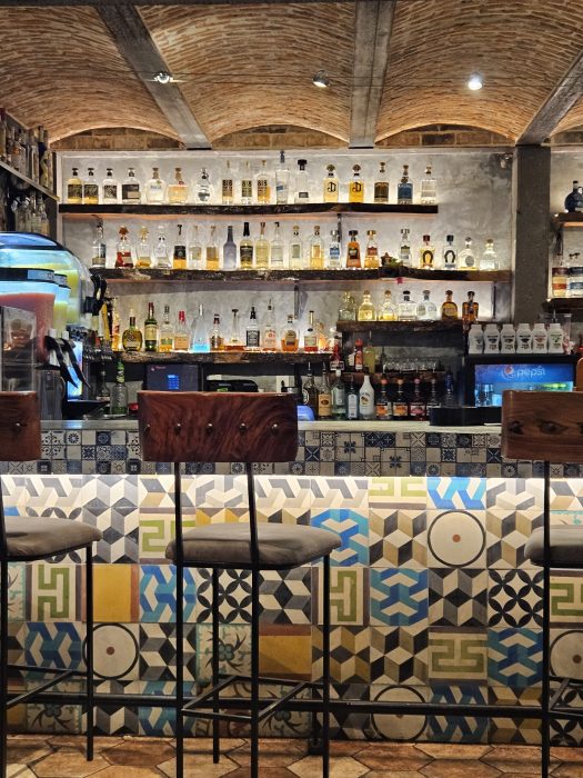 Bar interior at Cantina Del Sol with a patterned tile counter, three wooden barstools, and shelves displaying various bottles of liquor against a brick-arched ceiling.