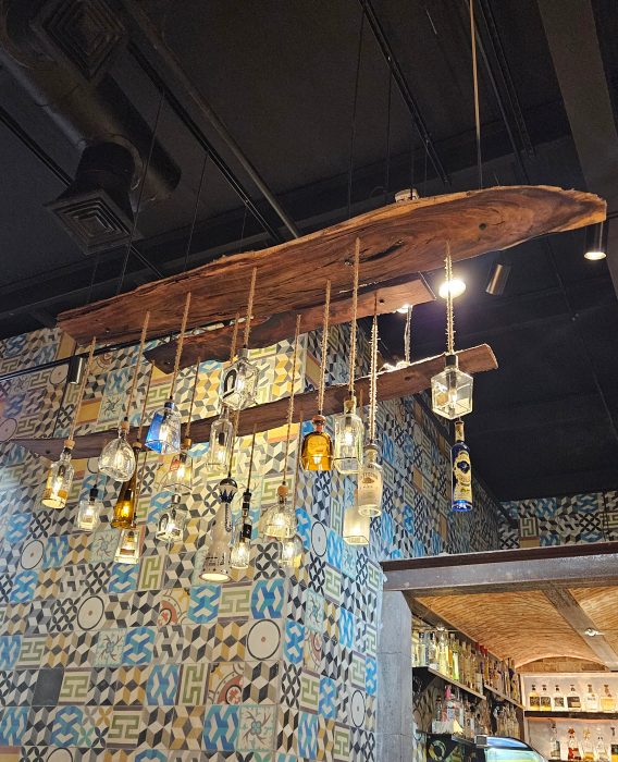 A ceiling light fixture from Cantina Del Sol, crafted with a wooden plank and hanging liquor bottles, stands out against a patterned tile wall in the background.