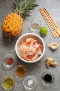 Ingredients for a shrimp dish are arranged on a gray surface: pineapple, shrimp, lime, garlic, ginger, skewers, and small bowls containing oil, soy sauce, salt, pepper, and chili flakes.