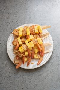 Skewers of shrimp and pineapple pieces are arranged on a white plate placed on a gray surface.