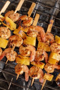 Grilled shrimp and pineapple skewers on a barbecue grill, showing char marks and placed over smoldering coals.