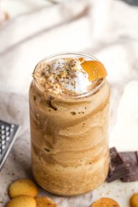A creamy, frothy coffee drink in a glass, topped with whipped cream, a peach slice, and a sprinkle of spice. Surrounded by chocolate pieces and small round cookies.