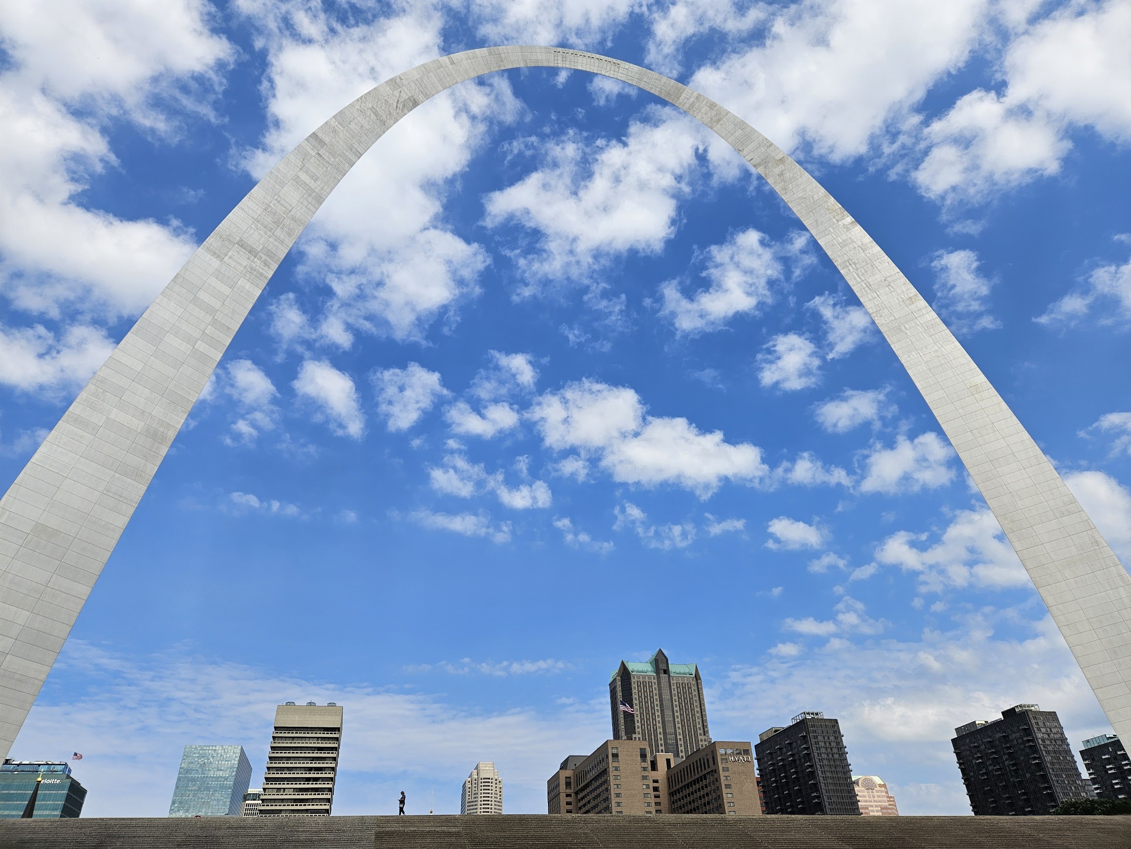 The Gateway Arch in St. Louis, Missouri, rises above city buildings under a partly cloudy blue sky.