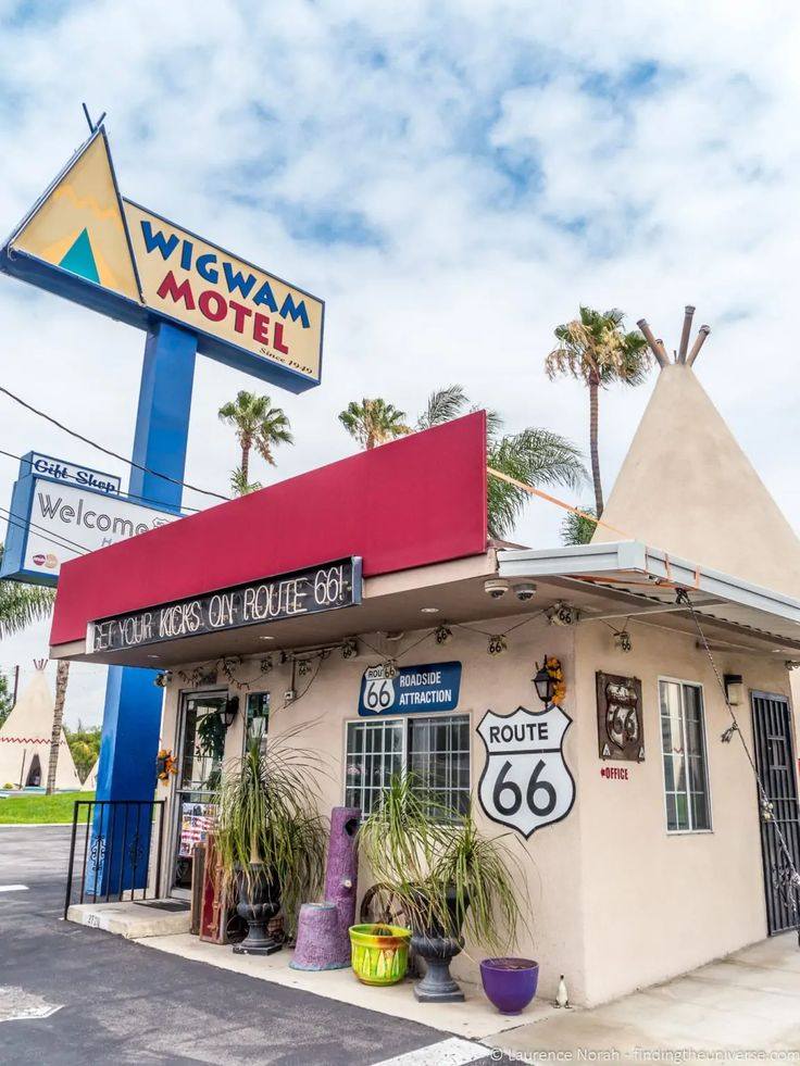 The Wigwam Motel on Route 66 features a small office building with tepee-style structures, a red awning, and a large roadside sign.