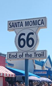 A Route 66 sign in Santa Monica reads “End of the Trail,” surrounded by crowds and nearby buildings, including the Playland Arcade.
