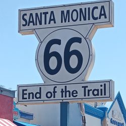 A Route 66 sign in Santa Monica reads “End of the Trail,” surrounded by crowds and nearby buildings, including the Playland Arcade.