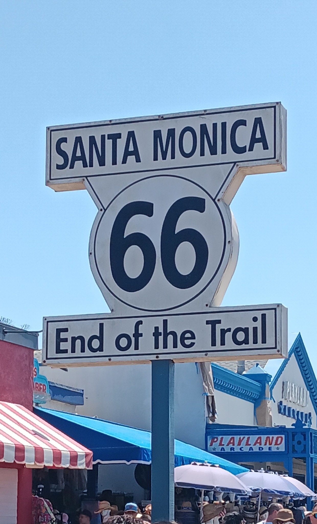 A Route 66 sign in Santa Monica reads “End of the Trail,” surrounded by crowds and nearby buildings, including the Playland Arcade.