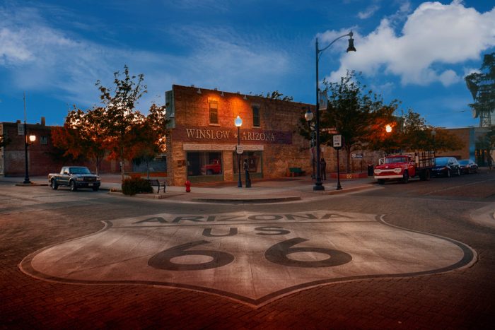 A street corner in Winslow, Arizona features a large Route 66 road emblem, classic cars, trees, and a brick building at dusk.
