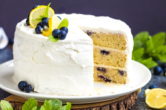 A three-layer lemon blueberry cake with white frosting, garnished with lemon slices, blueberries, and mint leaves, with one slice removed.