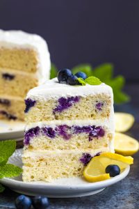 A slice of layered lemon blueberry cake with white frosting, garnished with blueberries, mint leaves, and lemon slices on a small plate.