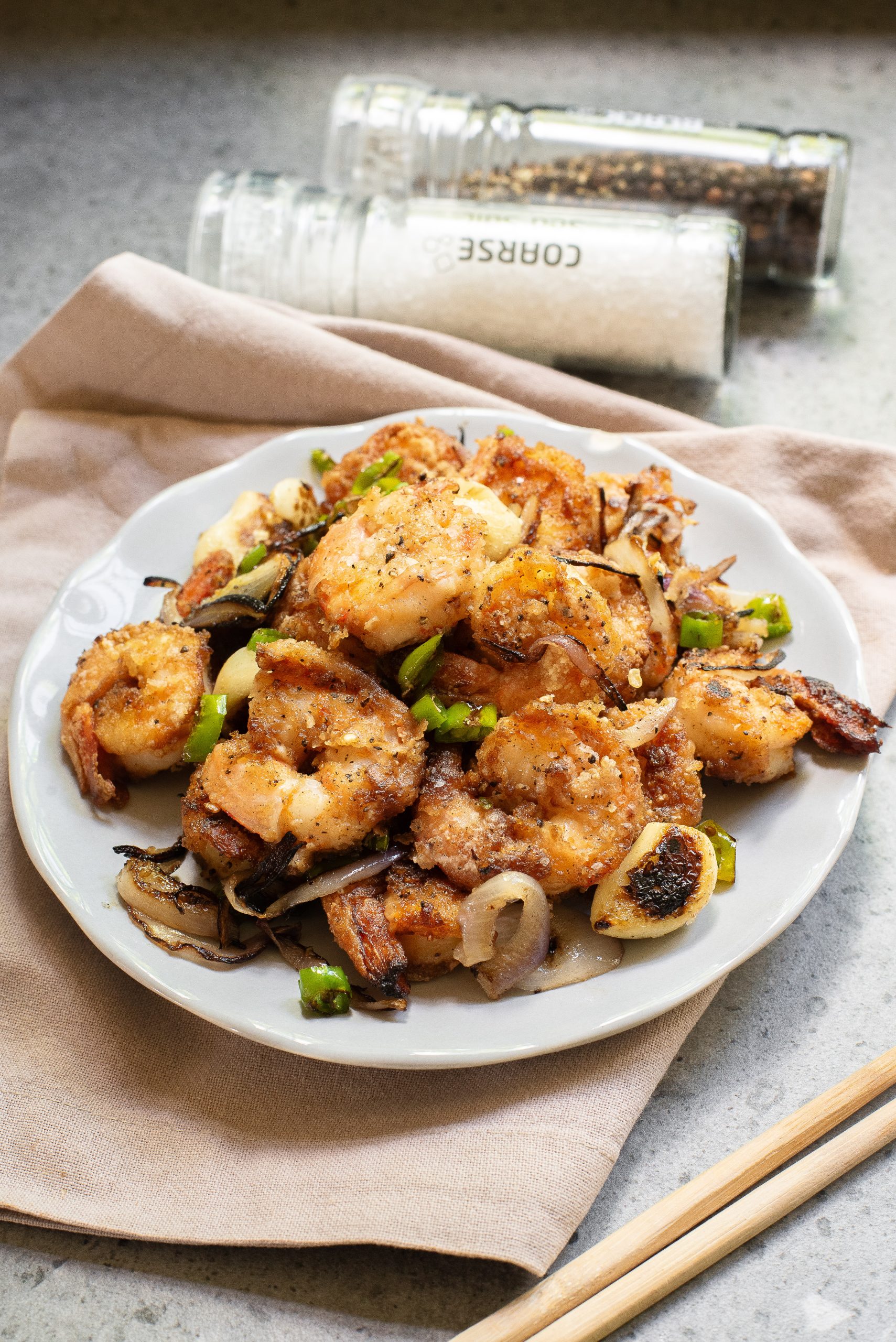 A plate of food with sliced onions and green peppers, placed on a beige napkin with chopsticks nearby and salt and pepper shakers in the background.