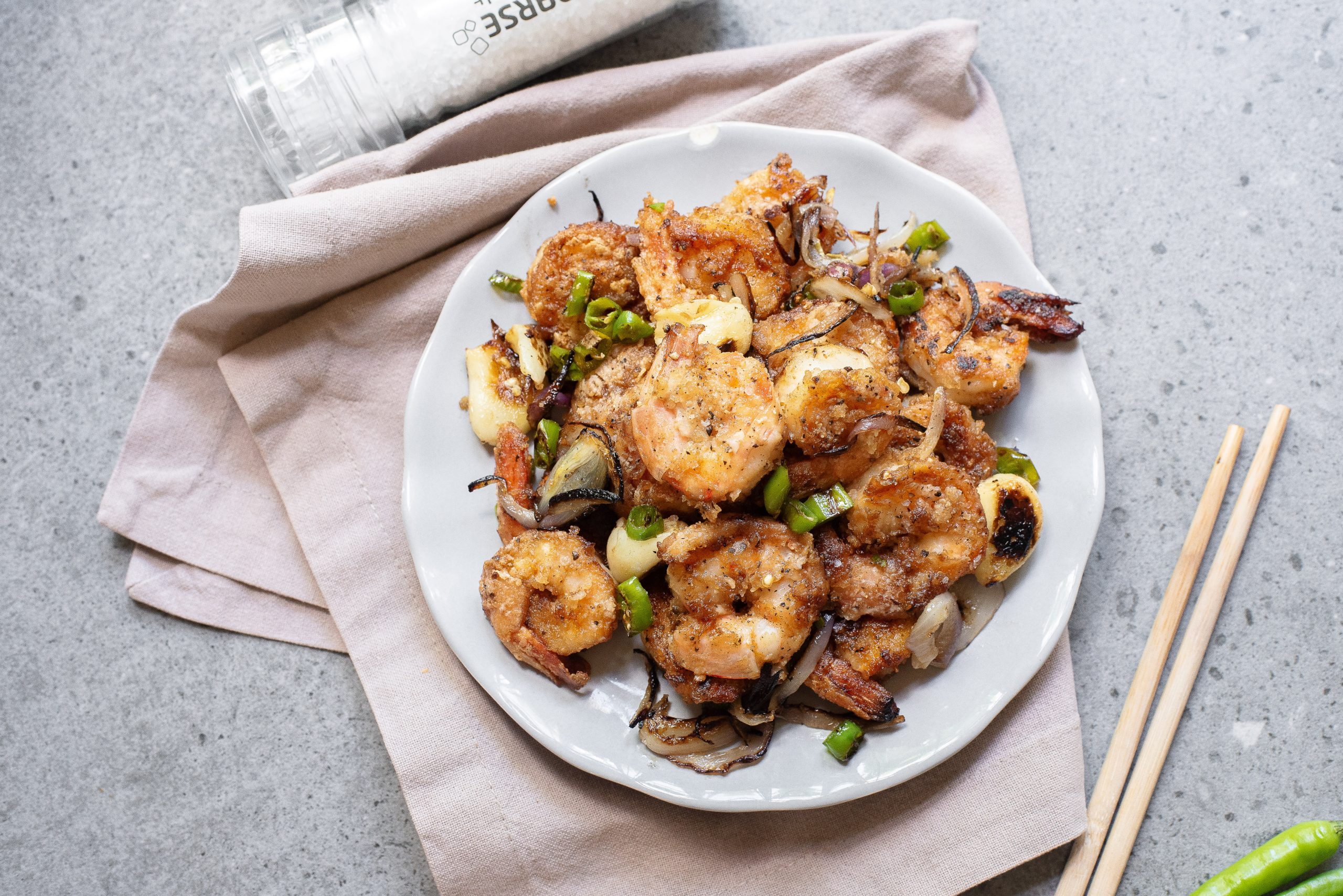 A white plate of cooked shrimp with sliced onions and green peppers sits on a beige napkin, next to chopsticks and a seasoning container on a gray countertop.