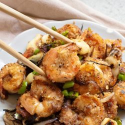 A plate of seasoned and sautรฉed shrimp with sliced onions and green peppers, with chopsticks picking up a shrimp.