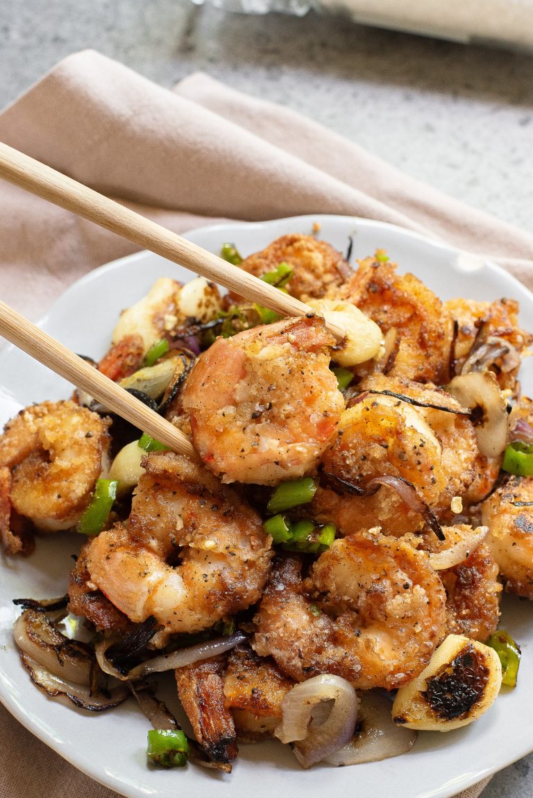 A plate of seasoned and sautéed shrimp with sliced onions and green peppers, with chopsticks picking up a shrimp.