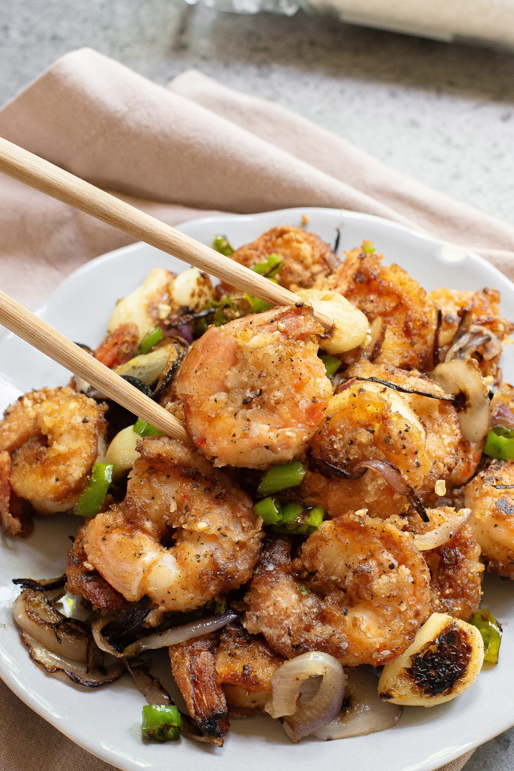 A plate of seasoned and sautรฉed shrimp with sliced onions and green peppers, with chopsticks picking up a shrimp.