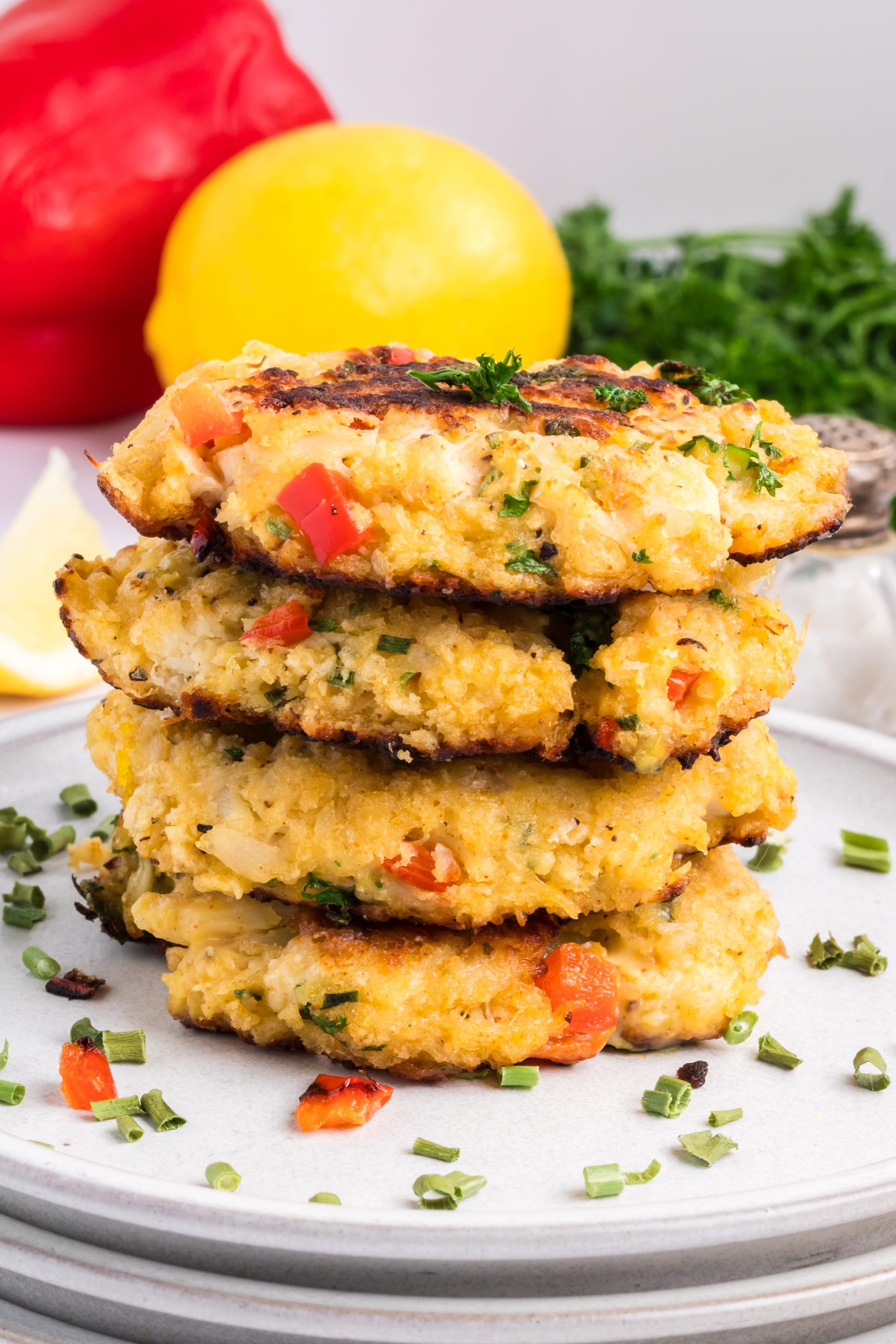 A stack of four Classic Crab Cakes with visible red and green peppers, served on a white plate with chopped herbs sprinkled around.