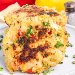 Two golden-brown crab cakes with chopped herbs and red pepper are stacked on a plate, with a lemon, red bell pepper, and salt shaker in the background.