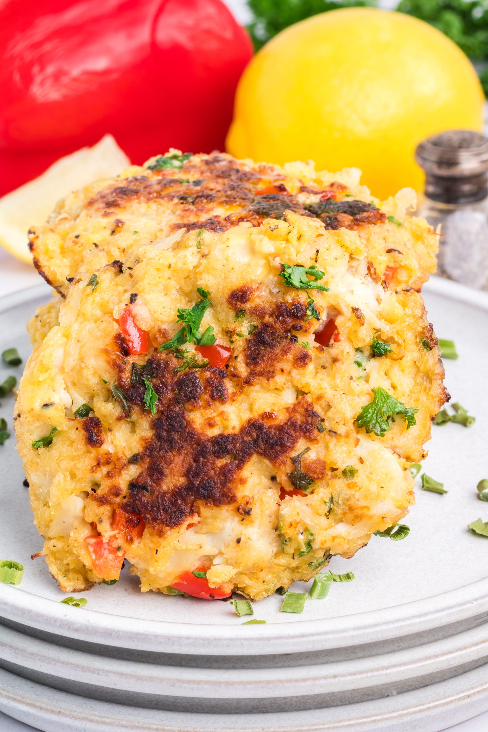 Two golden-brown crab cakes with chopped herbs and red pepper are stacked on a plate, with a lemon, red bell pepper, and salt shaker in the background.