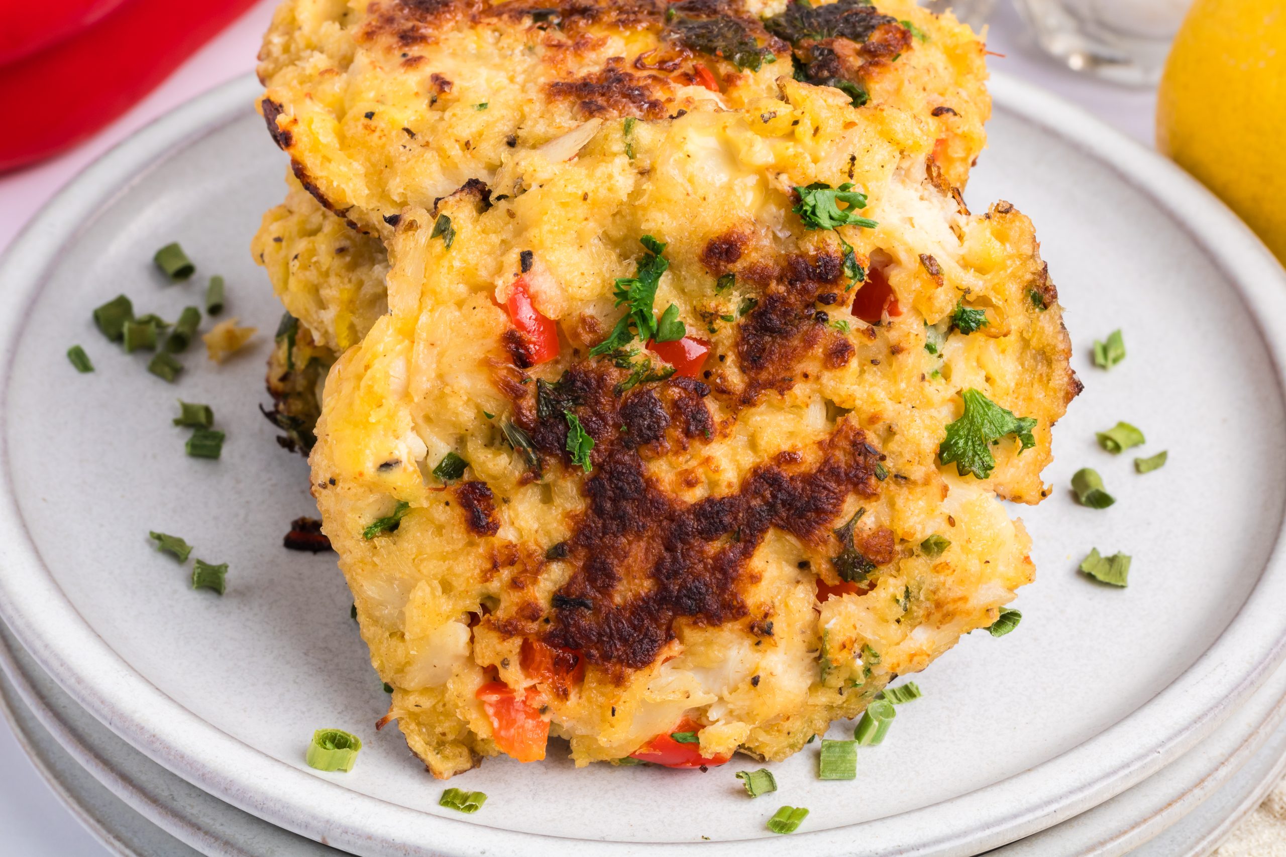 A close-up of two golden-brown crab cakes with visible herbs and red pepper pieces, served on a white plate.