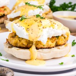 An English muffin topped with a crab cake, poached egg, and hollandaise sauce, garnished with parsley, on a white plate.
