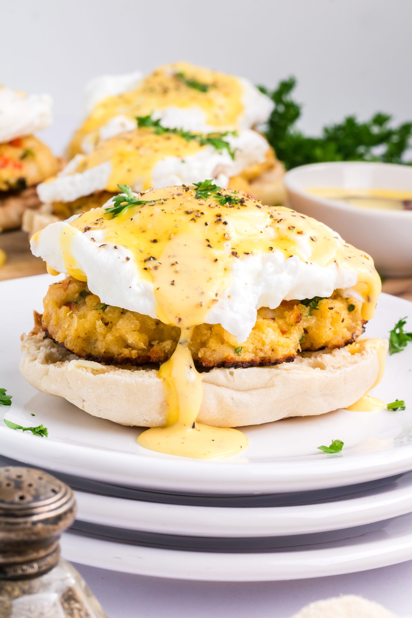 An English muffin topped with a crab cake, poached egg, and hollandaise sauce, garnished with parsley, on a white plate.
