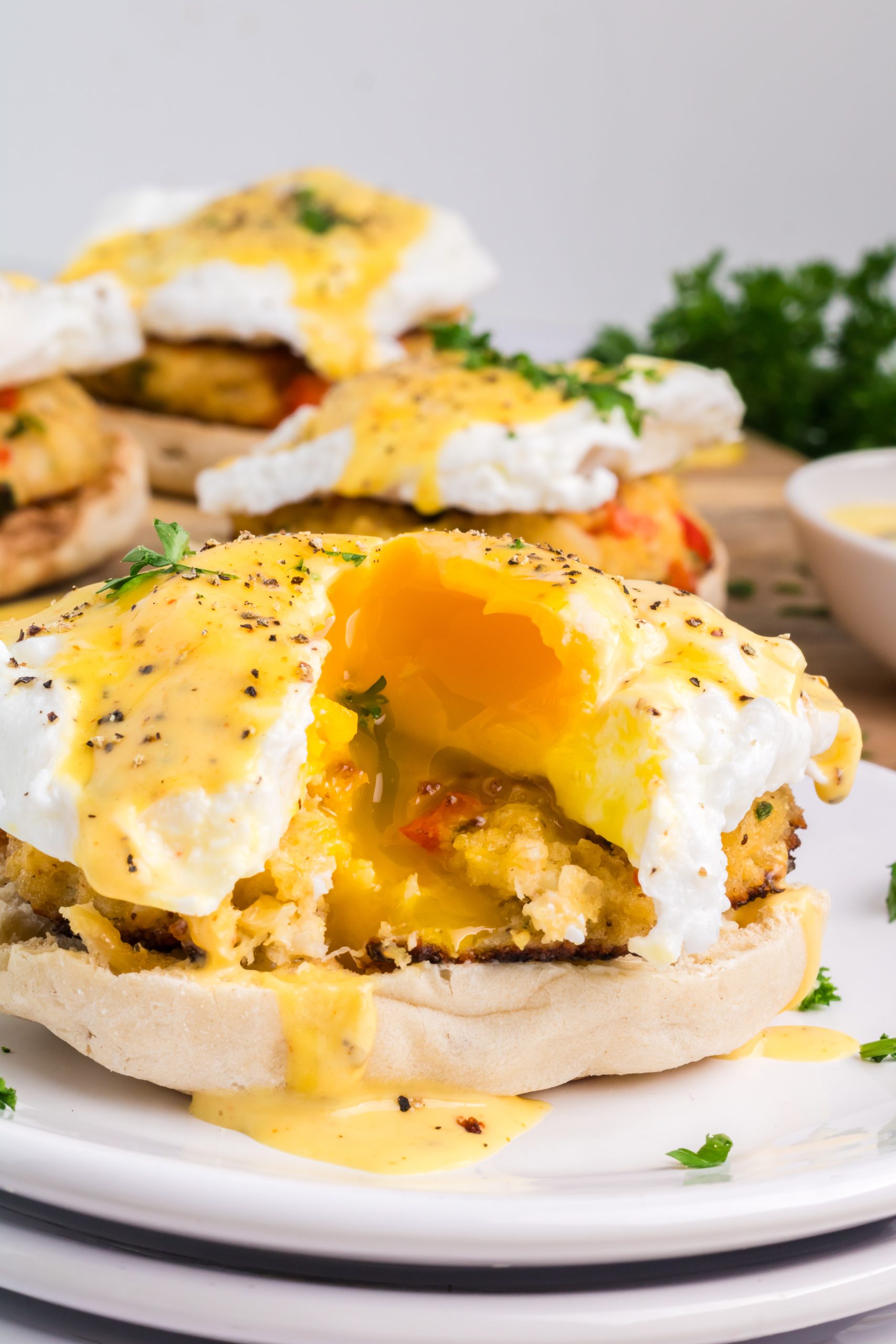 A poached egg on an English muffin with hollandaise sauce, served atop savory crab cakes and garnished with herbs and black pepper, with runny yolk spilling out.