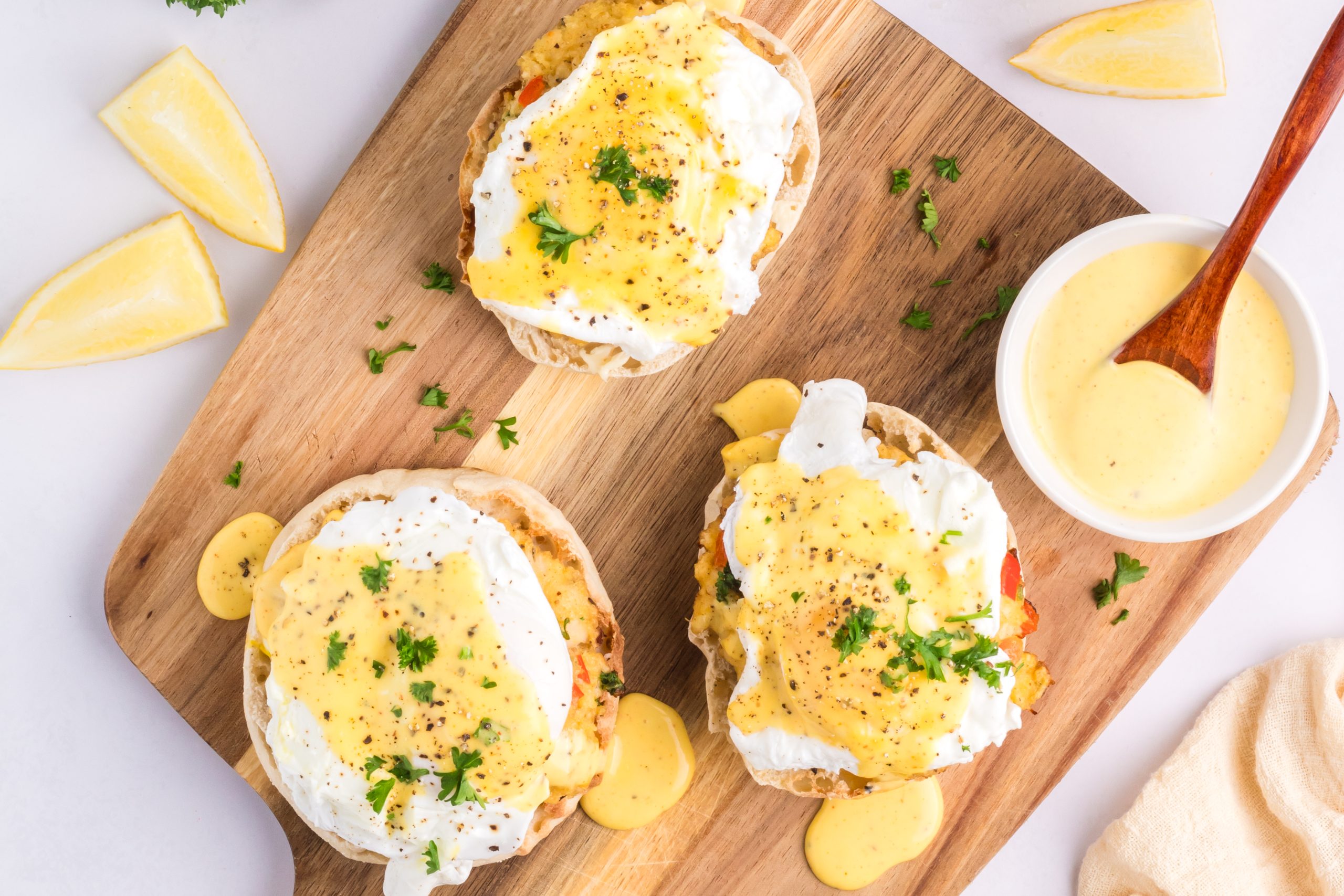 Three breakfast sandwiches served on a wooden board with a bowl of extra sauce and lemon wedges.