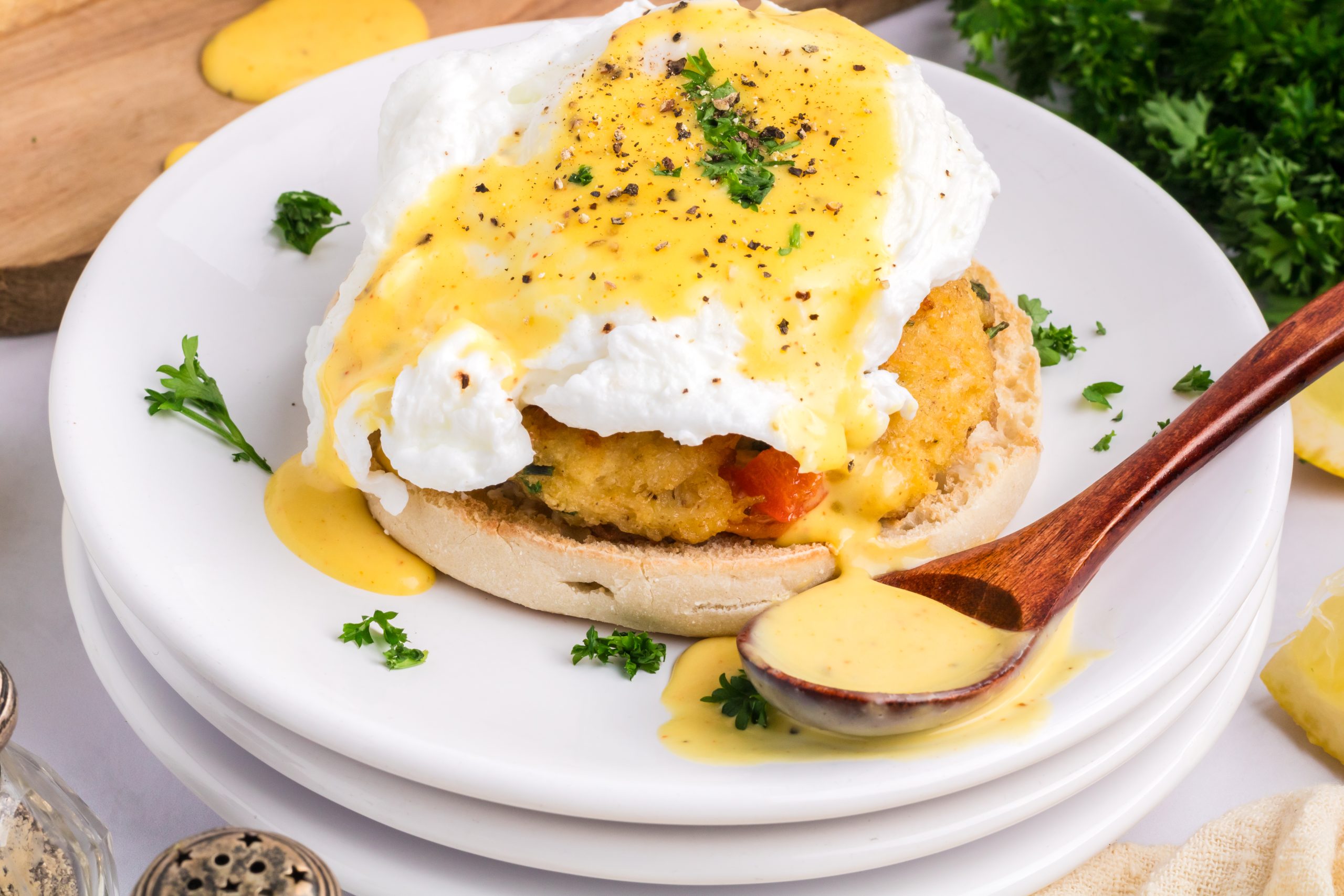 Eggs Benedict with a poached egg, hollandaise sauce, and a breaded patty on an English muffin, garnished with parsley and served on a white plate.