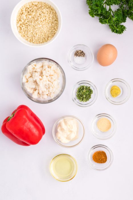 Top-down view of ingredients on a white surface, including red bell pepper, egg, herbs, spices, mayonnaise, oil, bread crumbs, parsley, and chopped white seafood.