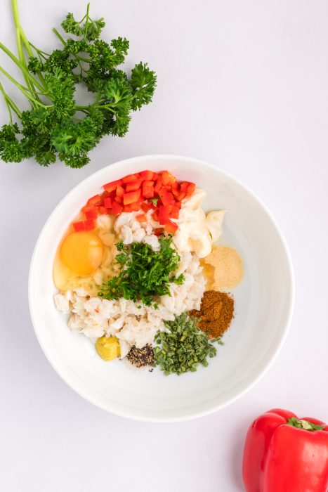 A white bowl containing chopped red pepper, egg, mayonnaise, crab meat, parsley, spices, and seasonings sits on a white surface with fresh parsley and a red bell pepper nearby.