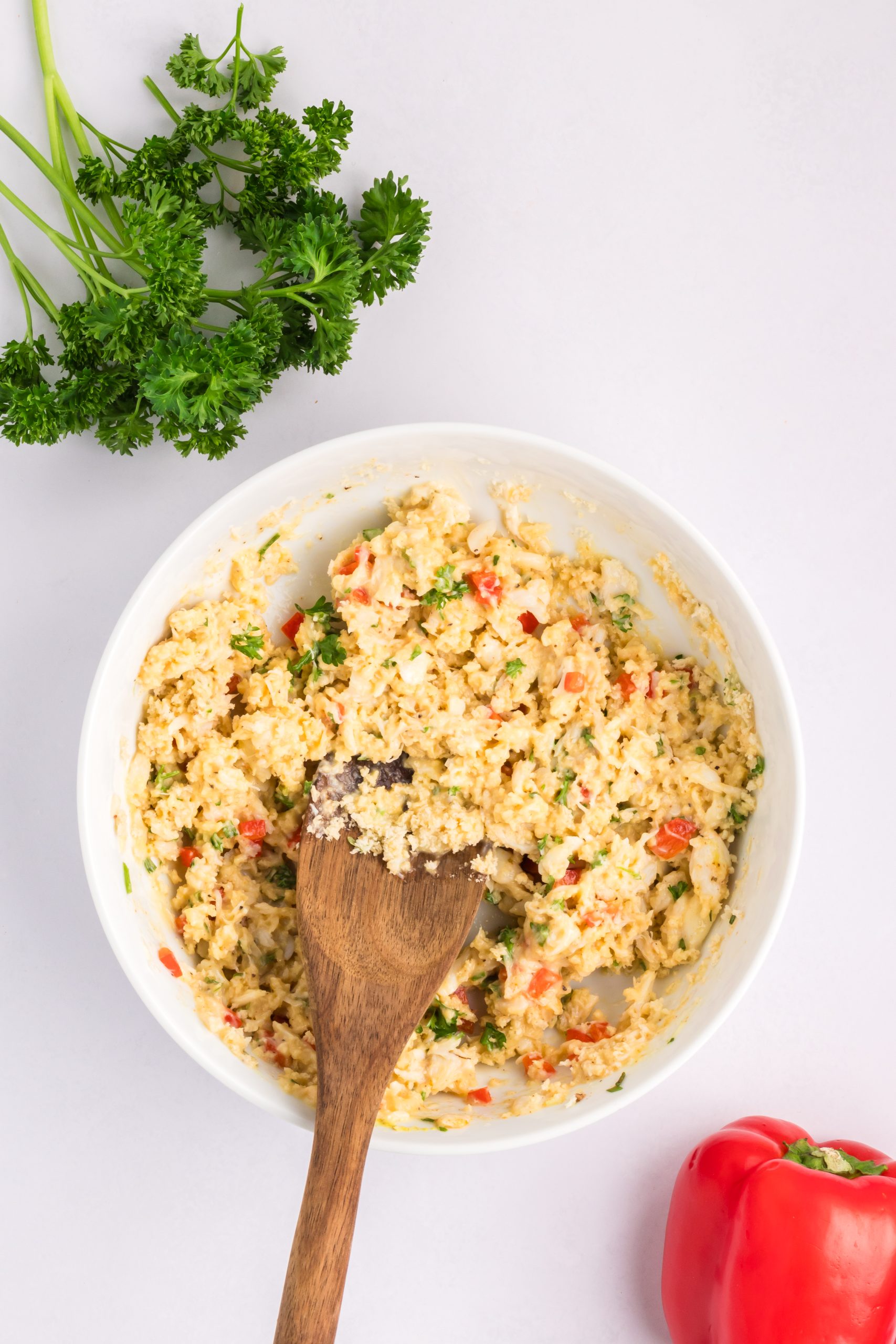 A bowl of mixed ingredients with chopped parsley and red bell pepper, stirred with a wooden spoon, sits on a white surface alongside fresh parsley and a red bell pepper.
