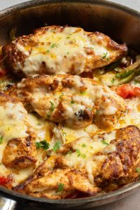 Cheesy baked chicken breasts topped with herbs and melted cheese in a skillet, surrounded by vegetables.