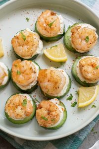 A plate of cucumber slices topped with a dollop of creamy spread and cooked shrimp, garnished with herbs and lemon wedges.
