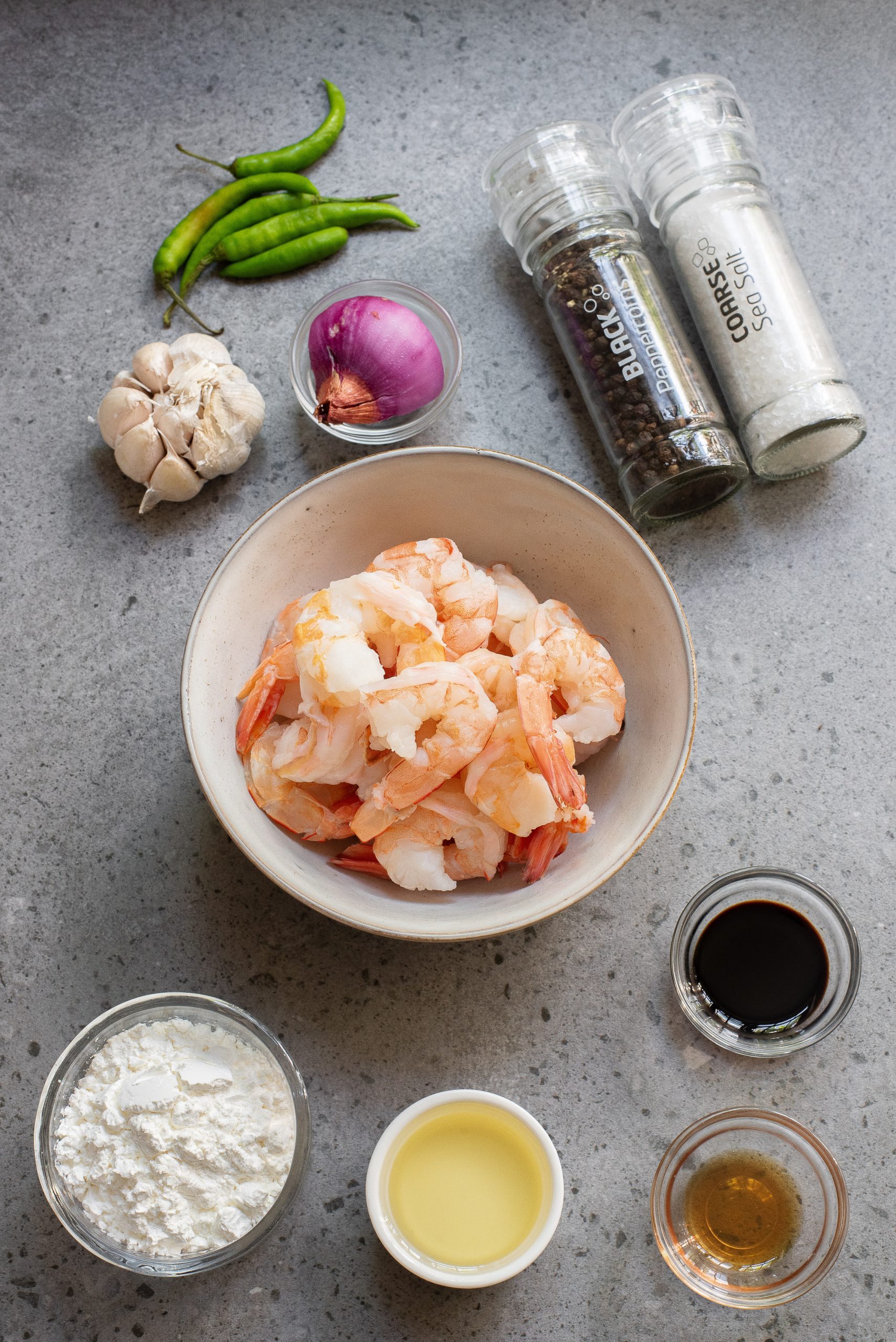 A bowl of peeled shrimp surrounded by green chilies, garlic, red onion, black pepper, salt, soy sauce, flour, cooking oil, and a small bowl of clear liquid on a grey surface.