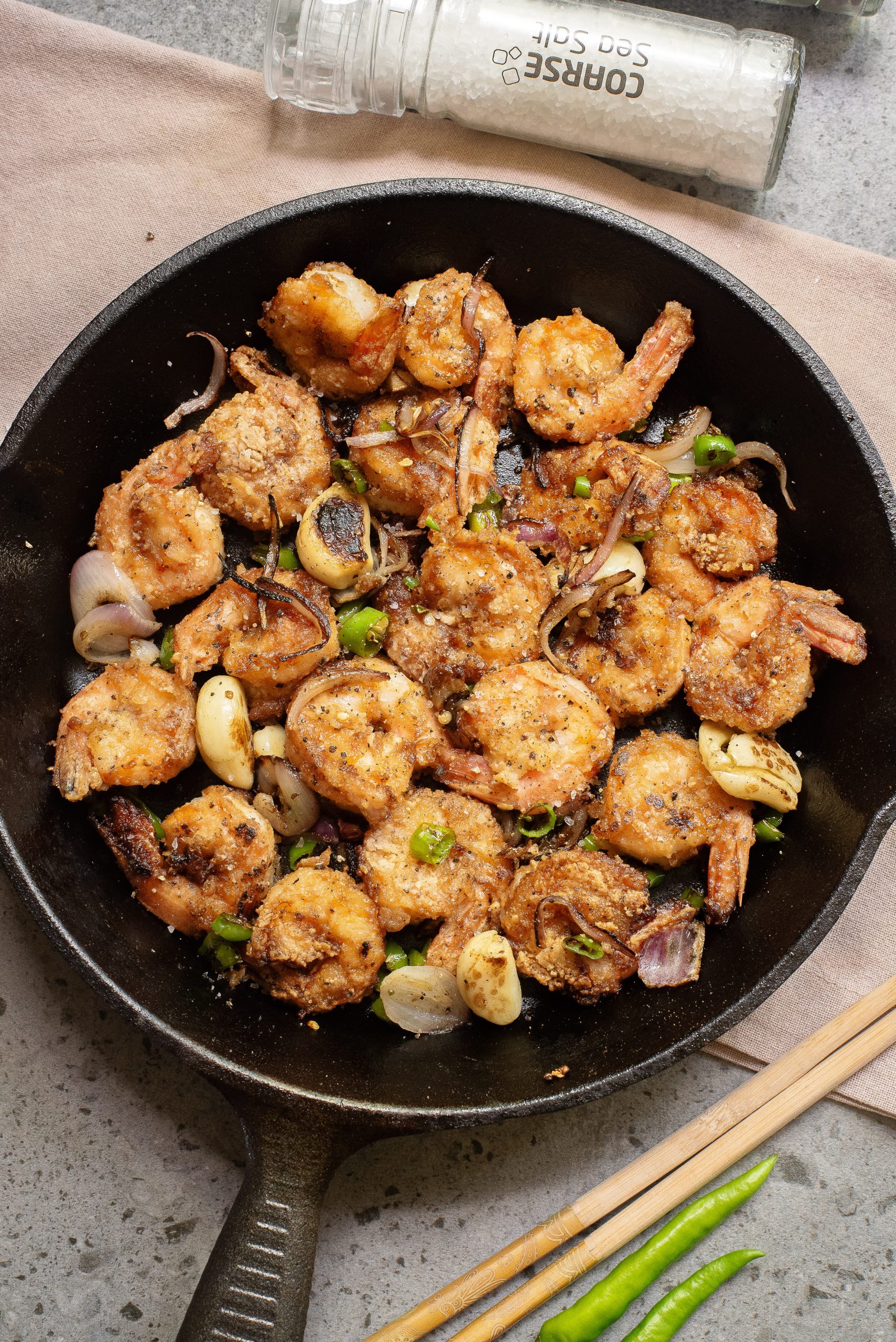 A cast iron skillet filled with cooked, seasoned seafood, sliced garlic, green chilies, and onions, with a coarse salt grinder in the background.