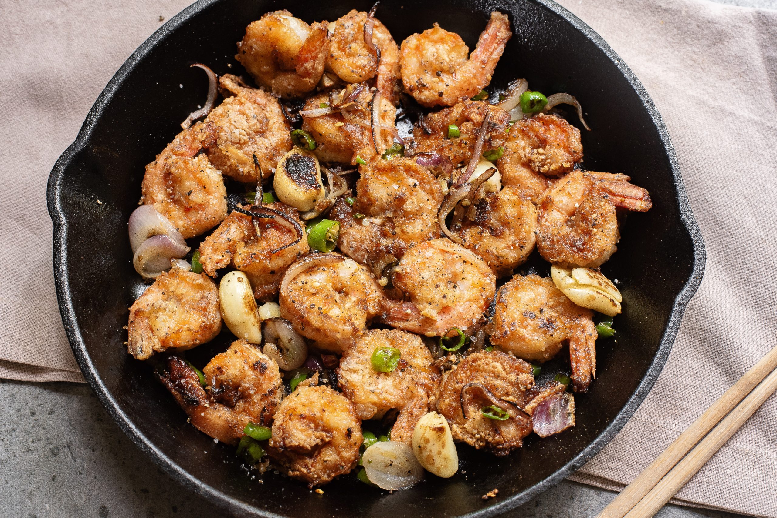 A cast iron skillet filled with cooked, breaded shrimp, garlic cloves, sliced onions, and green onions on a beige napkin.
